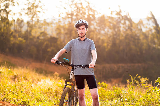 Portrait of a Brazilian cyclist with his mountain bike standing standing in the middle of a trail and the sunlight in the background coming through the trees.