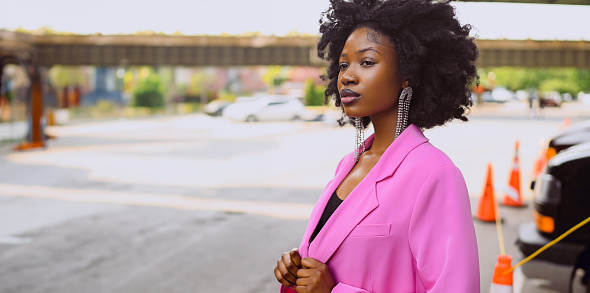 Fashion outdoor street style portrait of Beautiful young African American woman posing outside on urban city landscape summer day wearing pink jacket. Attractive black female, diversity concept