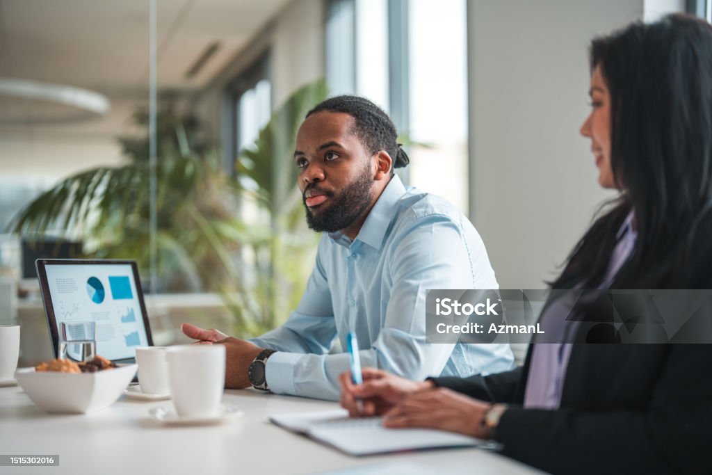 Mid Adult African Male Entrepreneur Reporting to Asian Female Branch Manager Mid adult African man showing project progress on laptop while sitting in a meeting room, his Asian female associate listening to him. Waist up image, coffee cups and snacks on the table top. Office Stock Photo