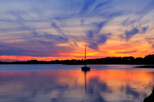 Sunset and sailboat in Martha's Vineyard, Massachusetts. Dramatic sky with vivid orange and blue cloudscape reflected on the water. This is a small salted lake forming a bay around the costline, connected to the sea.