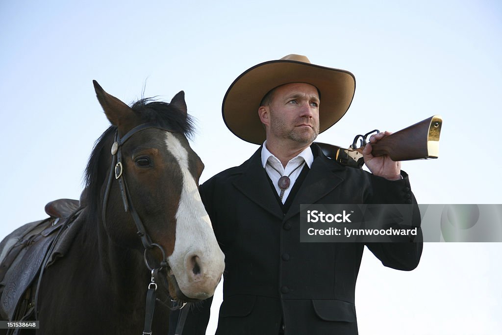 Man in fine old western clothes with horse & rifle Man gazing out with rifle over his shoulder and holding a saddled bay horse; blue sky behind. Blue Stock Photo