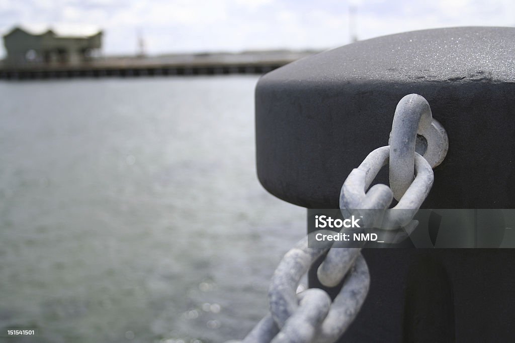 Concrete Piller with Chains by the Seaside Black Concrete Piller / Pilon by the Beach Seaside, with corroded steel chains over looking a blurred pier and hut in the background on an overcast day. Chain - Object Stock Photo