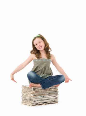 8 year old girl with long brown hair sitting cross legged on pile of newspapers for recycling. Please look at my portfolio for more images with this model against a white background.