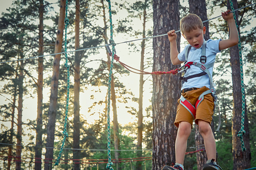 Brave boy carefully walks sideways on swinging rope, holding the rope above his head with his hands. In the children's rope park, child in equipment deftly and quickly passes difficult obstacle course