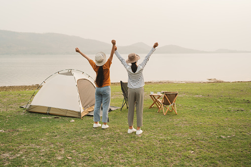 Back view. Asian two teenager girl raised up hand looking at river. Asia teen girl enjoyment happy smile amidst meadow field and mountain at sunset. Concept camping of lifestyle vacation and freedom