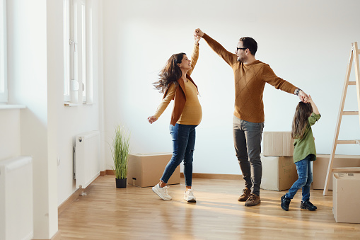 Happy pregnant family having fun while holding hands and dancing after relocating into a new home. Copy space.