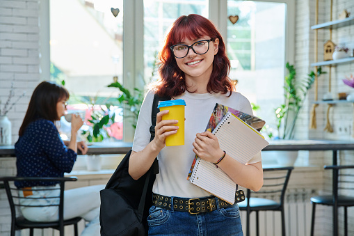 Portrait of young female student with takeaway coffee in college coffee shop. Girl 18-20 years old with backpack, looking at camera with textbooks, break for rest. Youth, leisure, lifestyle concept