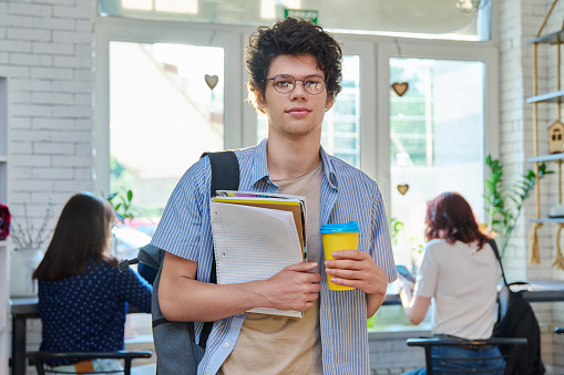 Portrait of young male student with takeaway coffee in college coffee shop. Guy 18-20 years old with backpack, looking at camera with textbooks, break for rest. Youth, leisure, lifestyle concept