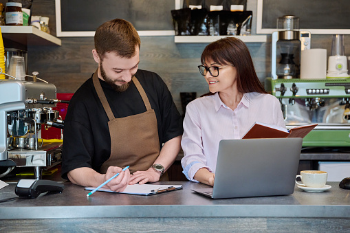 Colleagues partners, young male and mature woman talking working using laptop standing behind bar in coffee shop. Team, small business, work, staff, cafe cafeteria restaurant, entrepreneurship concept