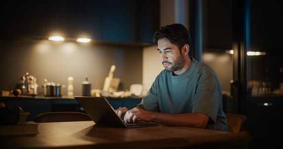Handsome Authentic Latin Man Sitting at a Table in a Cozy Kitchen and Typing on Laptop Computer at Home at Night. Male Adult Thinking While Browsing the Internet, Checking Videos on Social Networks.