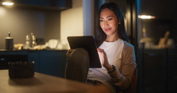 istock Beautiful Authentic Asian Woman Sitting at a Table in Cozy Kitchen and Using Digital Tablet at Home at Night. Female Smiling and Chatting on Social Media, Shopping Online, Browsing the Internet. 1516064684