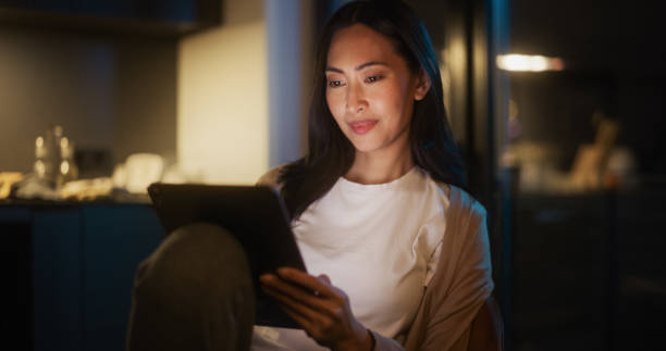 istock Beautiful Authentic Asian Woman Sitting at a Table in Cozy Kitchen and Using Digital Tablet at Home at Night. Female Smiling and Chatting on Social Media, Doing Online Shopping, Browsing the Internet 1516065221