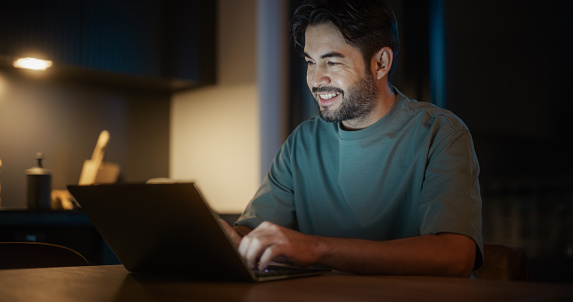 Handsome Authentic Latin Man Sitting at a Table in a Cozy Kitchen and Using Laptop Computer at Home at Night. Male Adult Smiling and Chatting on Social Media, Doing Online Shopping, Checking the News