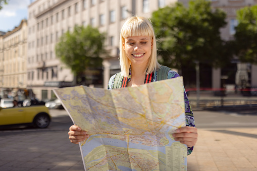 Young happy female tourist examining a map while standing on the city street.