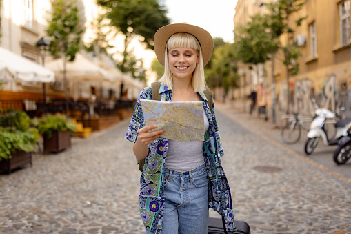 Happy female tourist examining a city map while pulling her luggage on the street.