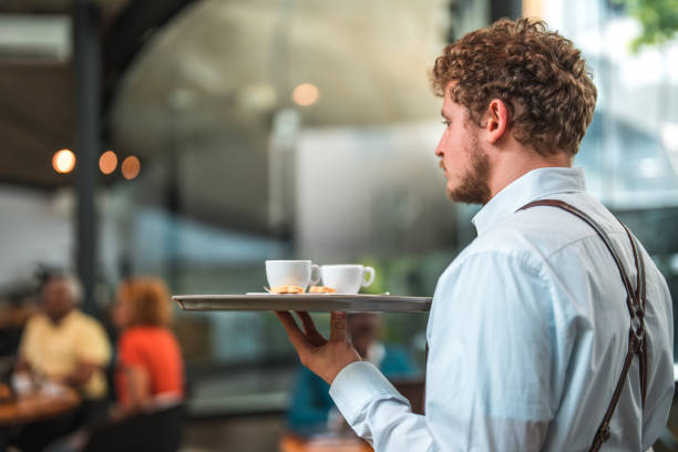 Appreciate the art of presentation as an accomplished Caucasian waiter, wearing a pristine shirt and apron, showcases his expertise by serving a tray adorned with meticulously crafted artisan coffee, adding a touch of sophistication to the modern cafe experience.