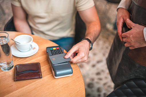 Close-up of hands of a Hispanic male embracing contactless payment with a credit card in a modern coffee house. The confident grip on the credit card showcases his seamless transition to contactless payment, effortlessly completing the transaction with a swift tap on the payment terminal.