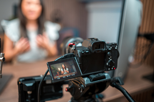 This striking photo features an Asian female content creator in a professional video recording setup. With a desktop PC and microphone, she confidently addresses the camera. The scene is elevated by meticulous lighting equipment placement.