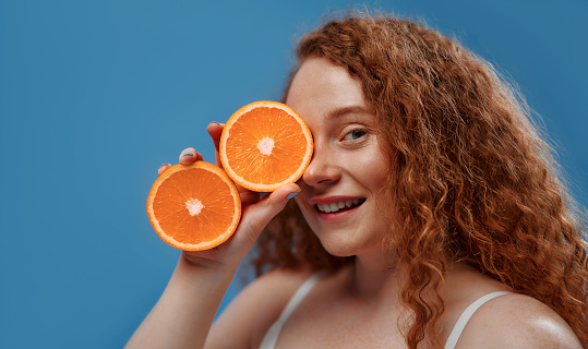 Freckled redhead plump plus size woman in white lingerie holding orange halves isolated on blue background. The concept of face and body skin care, body positive and cosmetology.