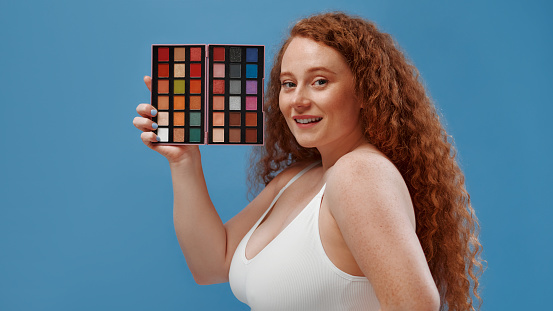 Beautiful plump plus size redhead woman with freckles holding eyeshadow palette isolated on blue background.