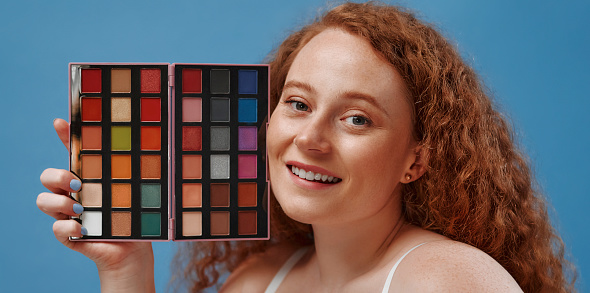 Beautiful plump plus size redhead woman with freckles holding eyeshadow palette isolated on blue background.