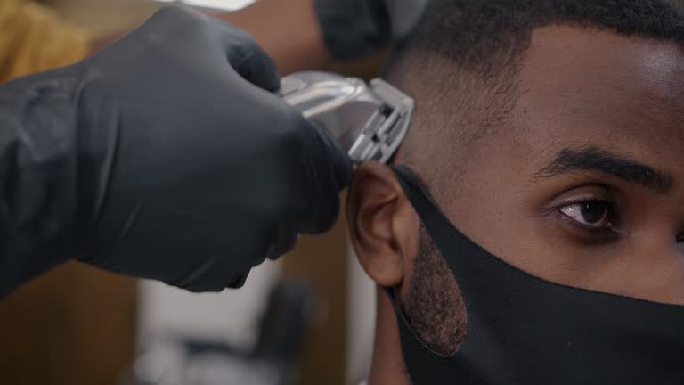 Close-up of hand in glove using shaving machine while man in medical face mask having haircut in barbershop