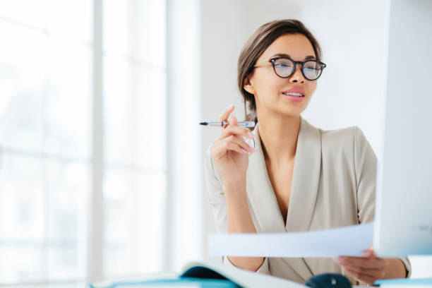 istock Focused businesswoman studies finances on computer, holds document and pen, works in office, wears formal attire and glasses 1516147468