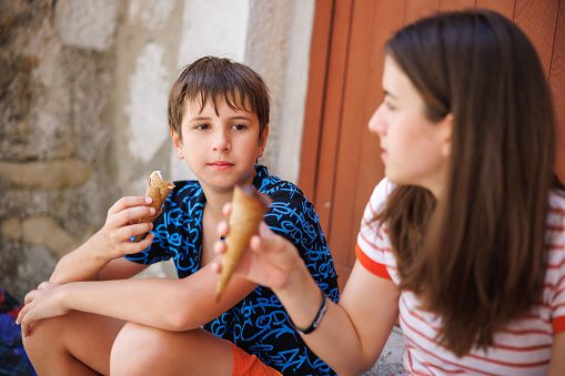Preadolescent boy and his teenage sister sitting on house doorway, talking and eating ice cream in cone, taking a break from town sightseeing on a summer day