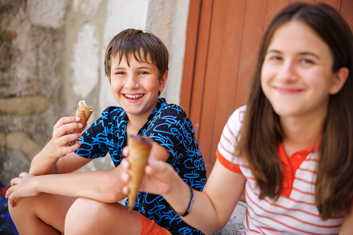 Preadolescent boy and his teenage sister sitting on house doorway, smiling and eating ice cream in cone, taking a break from town sightseeing on a summer day