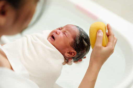 newborn baby girl crying while mother is washing her hair with sponge in a bathtub