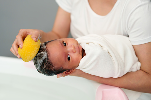mother is washing newborn baby hair with sponge in a bathtub