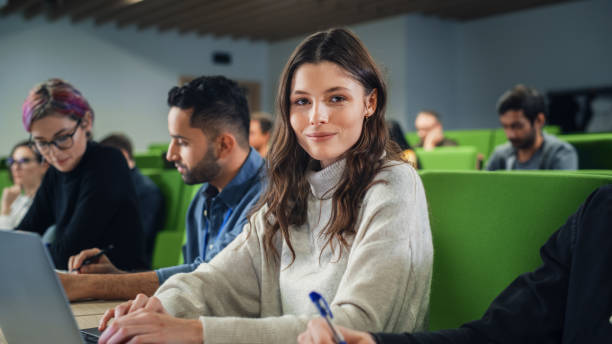 istock Beautiful Smart Female Student Studying in University with Diverse Multiethnic Classmates. Portrait of a Happy Caucasian Female Looking at Camera and Smiling. She is Using a Laptop Computer 1516269592
