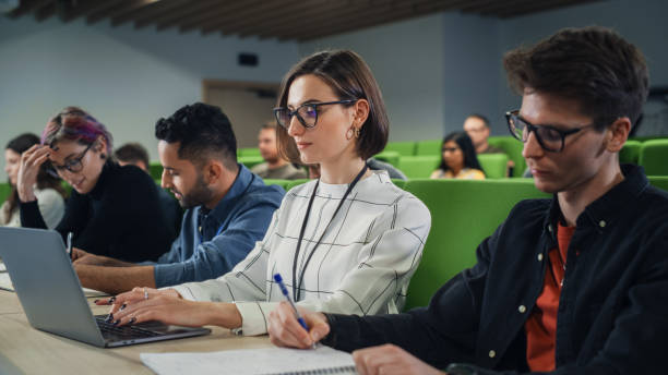 istock Young Female Student Studying in University with Diverse Multiethnic Classmates. She is Focused and Using a Laptop Computer. Patiently Listening to Professor's Lecture and Taking Notes 1516270123