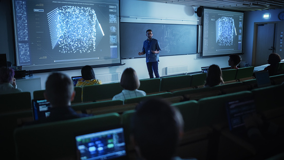 Young University Professor Explaining the Importance of Artificial Intelligence to a Group of Diverse Multiethnic Students in a Dark Auditorium. Teacher Showing Neural Network on Two Big Screens