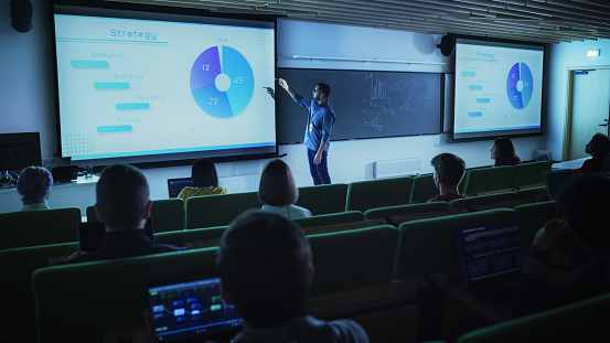 Young Male Teacher Giving a Business Strategy Lecture to Diverse Multiethnic Group of Female and Male Students in Dark College Room. Projecting Slideshow with Comparison Charts and Commercial Data