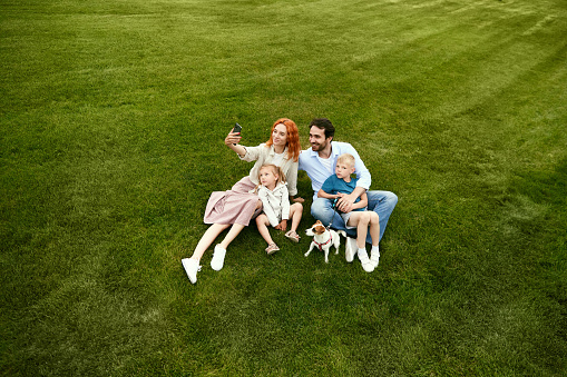 Young family, man and woman walking with their children in park, sitting on grass and taking selfie, photo together on warm summer day. Concept of family, childhood and parenthood, fun, weekends, love