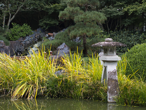 Japanese lantern stone ornament sits on the edge of a still pond is strategically placed amongst rocks, boulders and plants to create a peaceful environment.