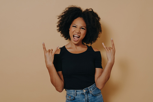 Young afro-american rock star, hands in rock sign, shouting with emotion, carefree, against beige background