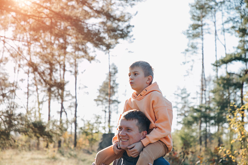 Dad and son on a walk in the forest in sunny weather. Family leisure. The father carries his son on his shoulders. Front view