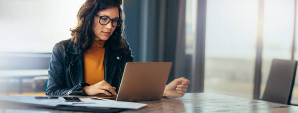 Asian business woman working on a laptop in a professional office Asian business woman sitting at a table in an office, typing on her laptop with focus. Young female professional showing a dedication and commitment to her project at work. one person stock pictures, royalty-free photos & images