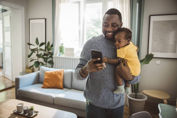 Father holding his baby Young Afro-american father at home holding his cute baby son in his arms, and using smart phone. father holding son stock pictures, royalty-free photos & images