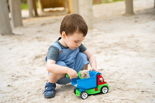 Little cute toddler boy 2.5 years old plays in the sandbox on a sunny summer day. Outdoor creative activities for kids. Toy car and shovel.