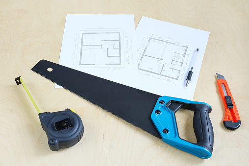 Sharp hacksaw, tape measure and knife on wooden surface, building tools next to blueprint of house
