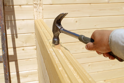 Hand holds hammer and strikes wood dowel to secure connection of wall timbers, building a new wooden house