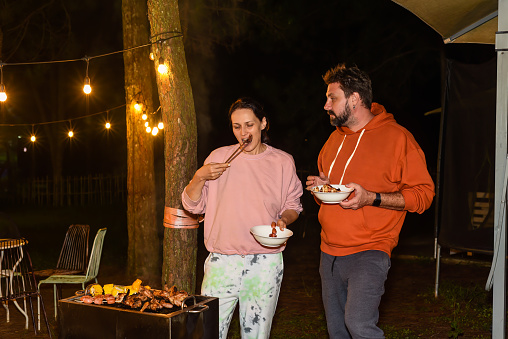 Pareja casada hombre y mujer comiendo salchichas de barbacoa por la noche cerca de guirnaldas de rayos en el patio trasero photo