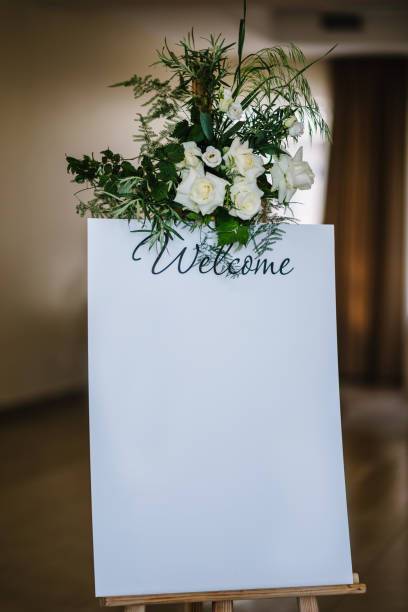 istock Welcome wedding. Space, place for text for seating guests on easel at reception. List, plan, chart seating area of guests at tables at restaurant. Frame is decorated with white flowers and greenery. 1516560763