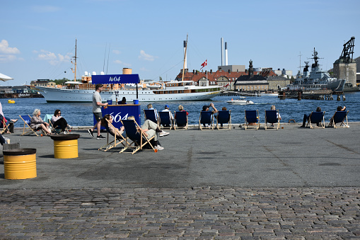Copenhagen, Denmark - May 14, 2023: Building Exterior, Copenhagen Harbor, Docked Danish Warship, Recreational Boat, People Sitting Down, Enjoying Beautiful Springtime Weather
