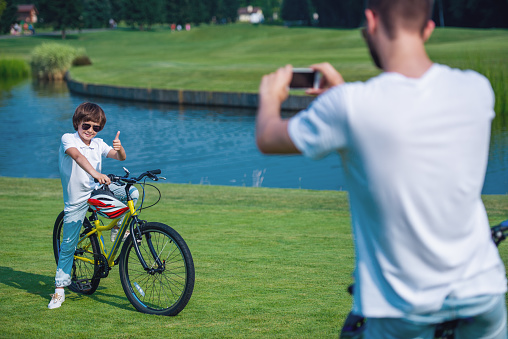 Father is taking photo of his son using a smartphone. Little boy is showing Ok sign, looking at camera and smiling while sitting on bicycle in park