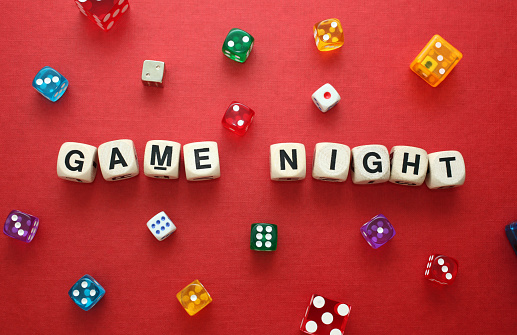 Close up photo with word dice showing the phrase 'Game Night', with a selection of colourful dice around it on a warm red cardboard backing.