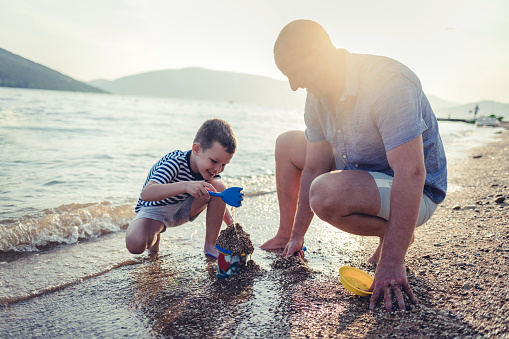 Photo of a little boy playing in the sand with his father, fully equipped with a beach supplies.